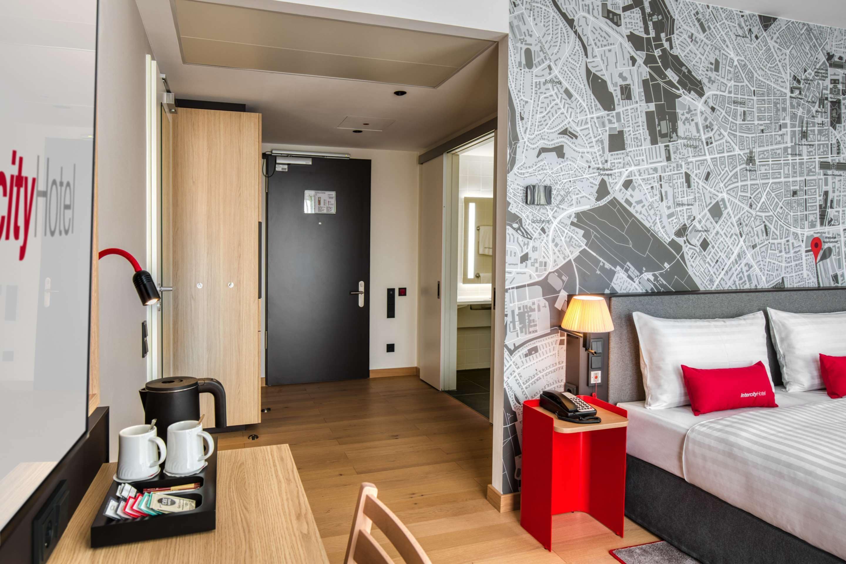 Habitación de hotel con una cama, una mesita de noche roja, una lámpara y una bandeja con tazas y un hervidor.