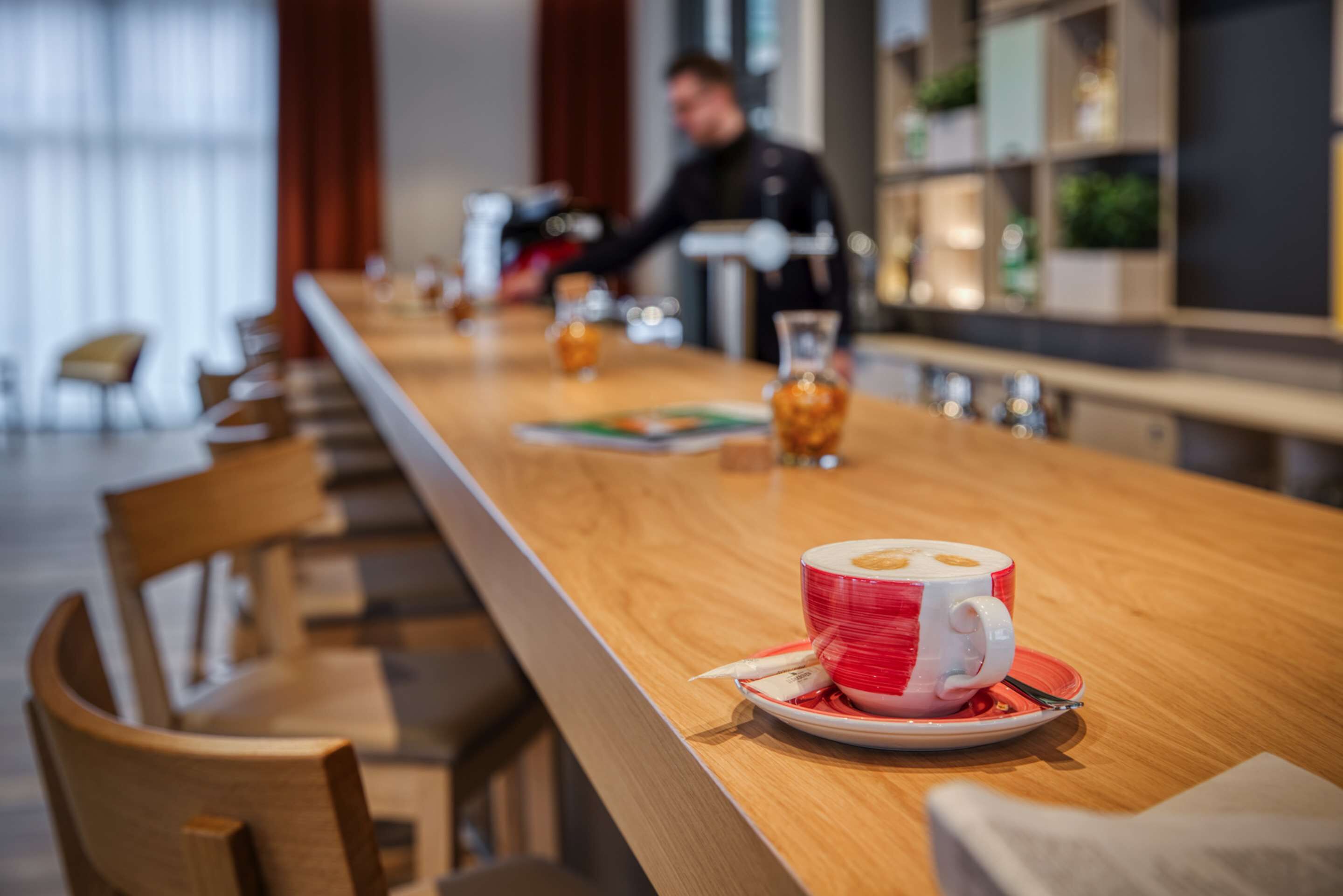 Una larga mesa de madera con una taza roja y blanca sobre un platillo, un vaso de líquido ámbar y un menú.