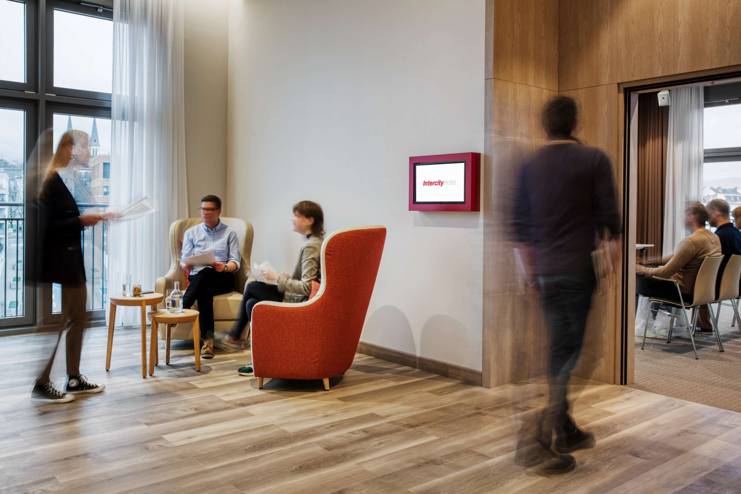 Un moderno vestíbulo de hotel con piso de madera, un sillón rojo y un muro de vidrio con vista a la ciudad.