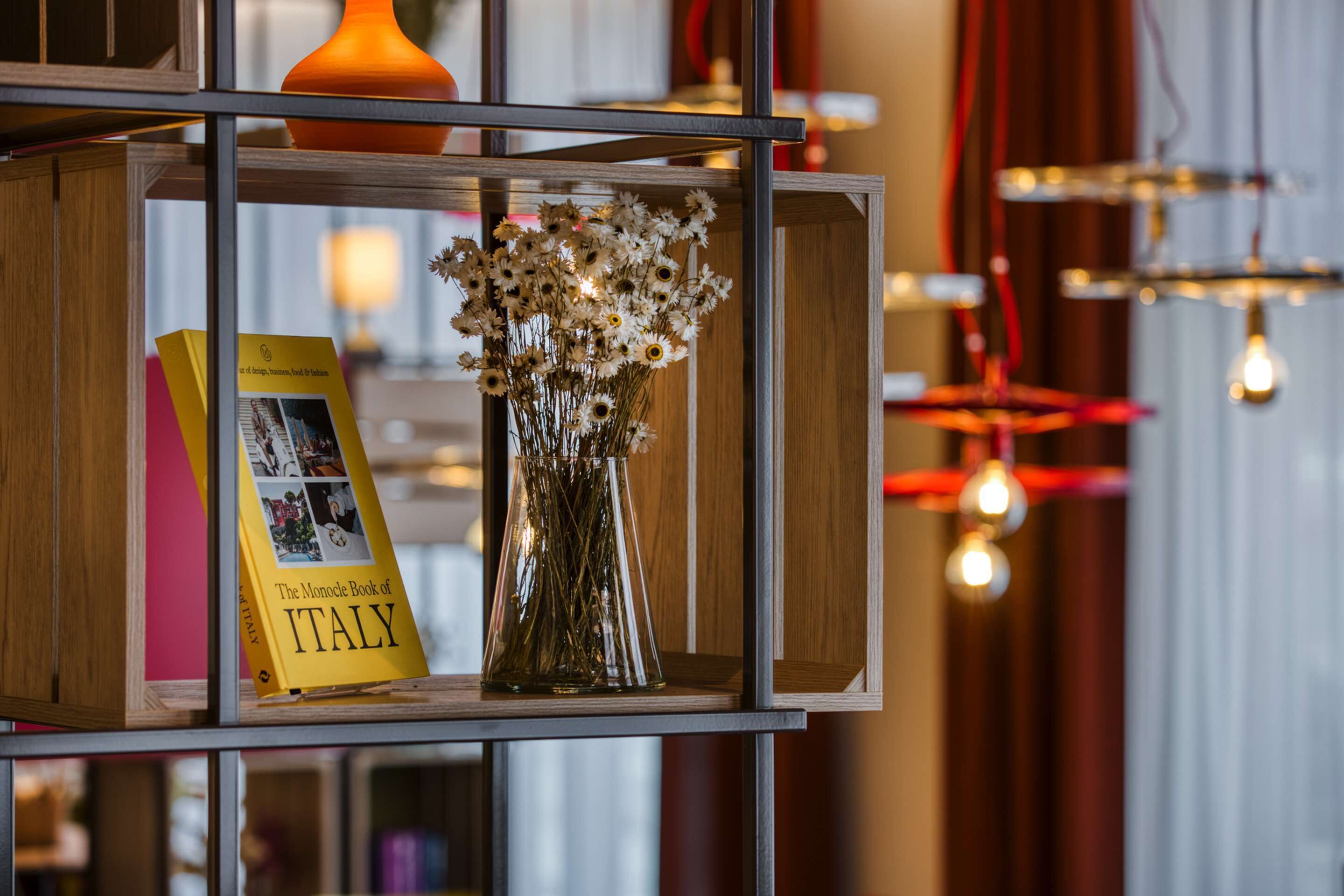 A shelf with a book titled 'The Monocle Book of Italy', a vase with dried flowers, and a hanging light fixture.