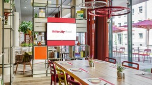 A modern hotel lobby with a large wooden table, colorful chairs, and a large screen displaying the hotel name. - IntercityHotel Wiesbaden (Wiesbaden)