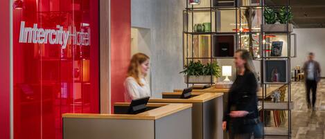 A hotel reception area with a red facade, a reception desk, and a shelving unit with plants and decorative items.