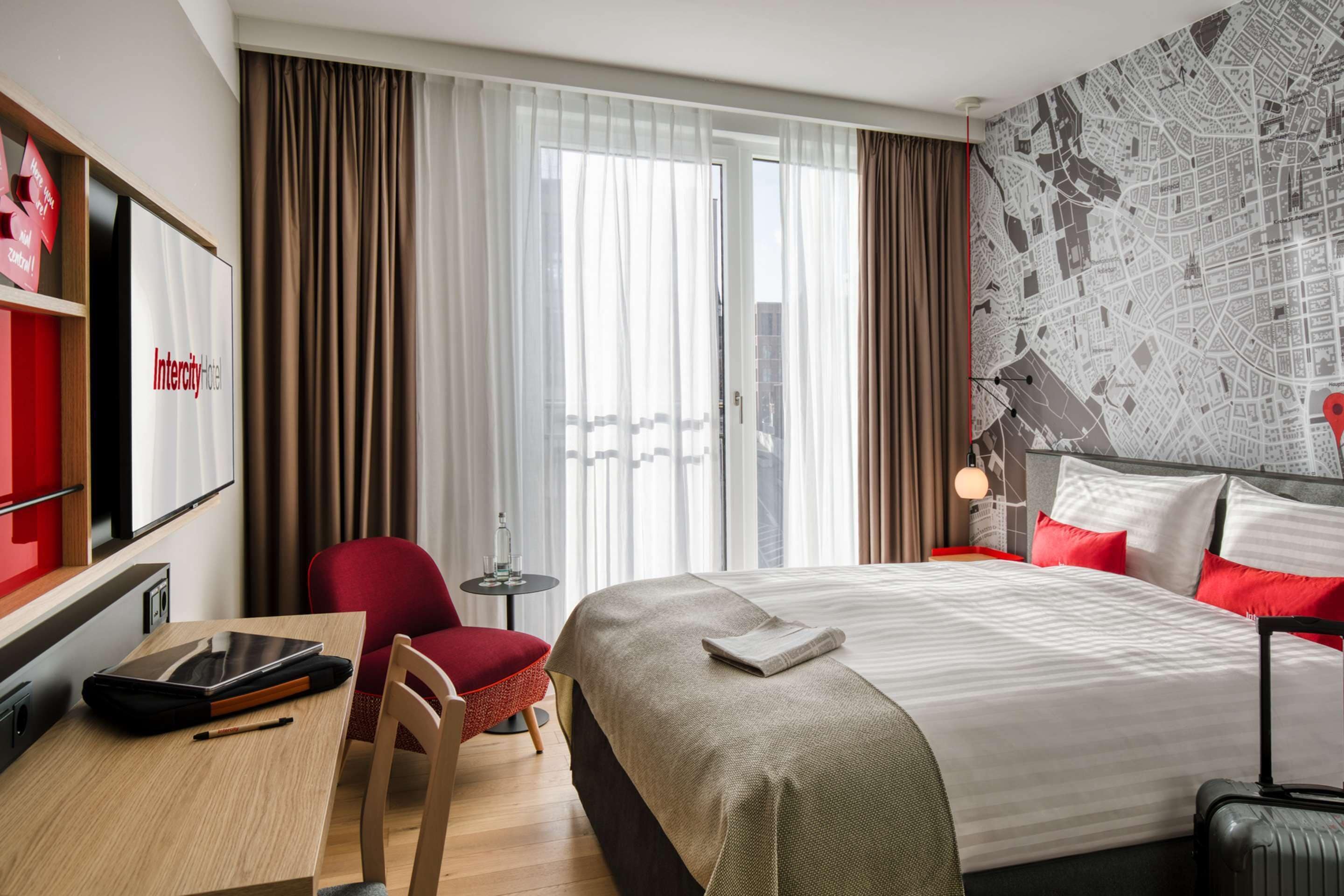 Ein Hotelzimmer mit Bett, Schreibtisch mit Tablet, Sessel, rotes Armlehnsofa, kleiner Tisch mit Lampe und Koffer.