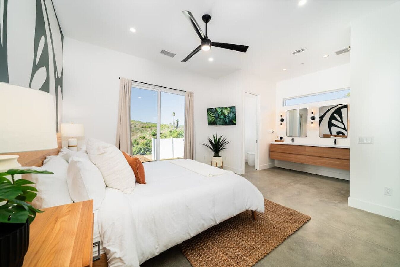 A modern bedroom with a large bed, a ceiling fan, a sliding glass door to a balcony, a bathroom with a vanity and mirror, and a view of greenery outside.