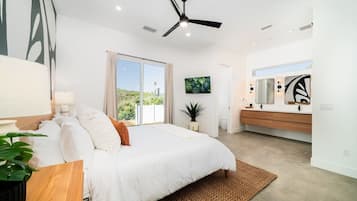 A modern bedroom with a large bed, a ceiling fan, a sliding glass door to a balcony, a bathroom with a vanity and mirror, and a view of greenery outside.