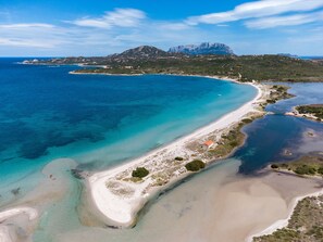 An aerial view of a coastal area with a beach, clear blue waters, and a mountain in the background.