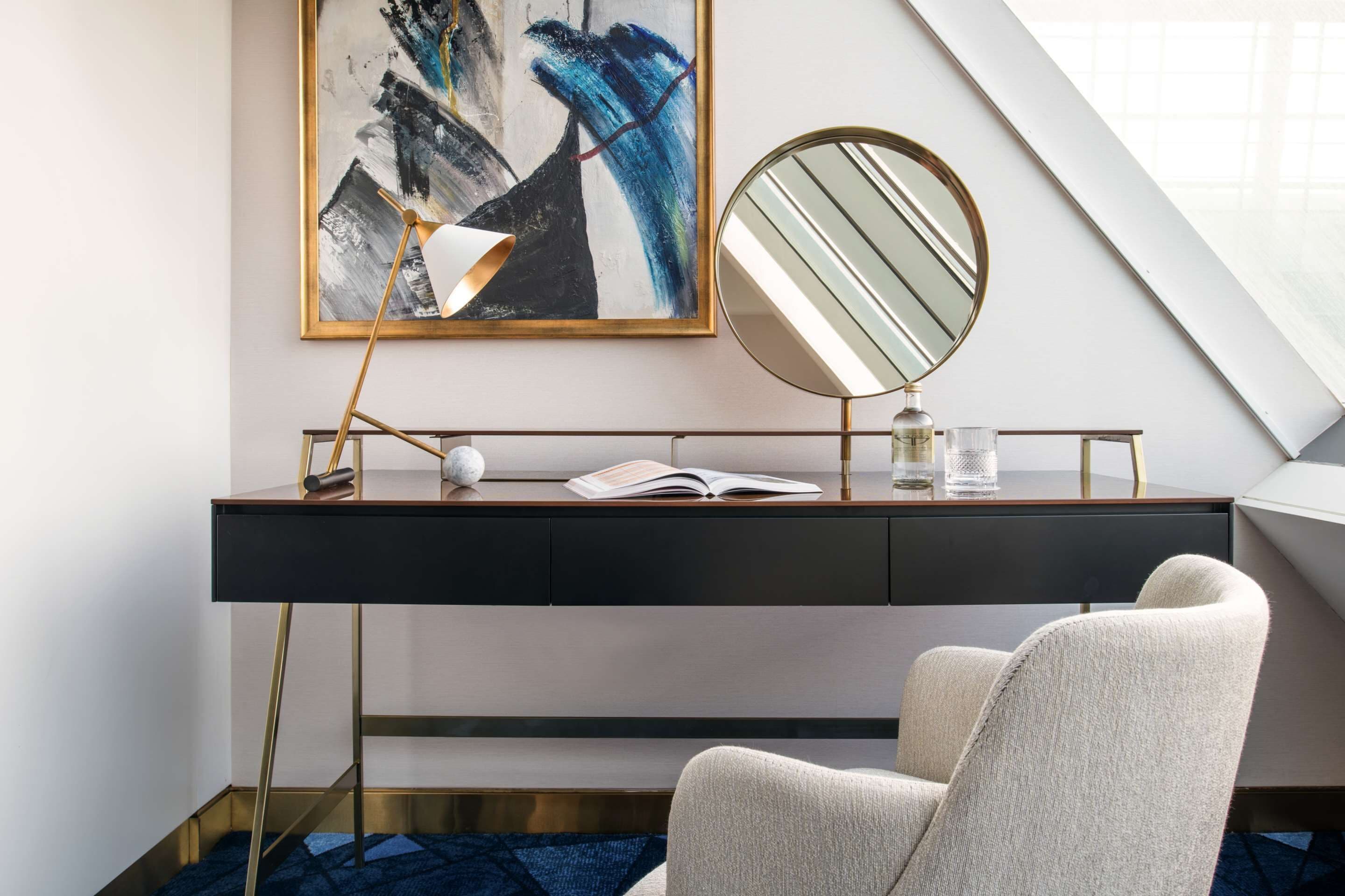 A modern hotel room with a desk, a round mirror, a painting, and a comfortable chair.