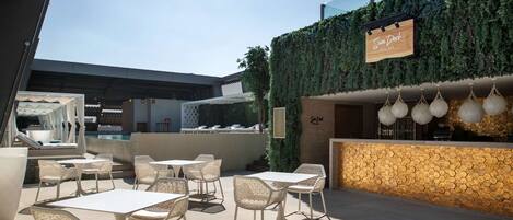 An outdoor patio area with white tables and chairs, a pool bar named 'Sun Dock', and a stone wall with hanging lanterns.