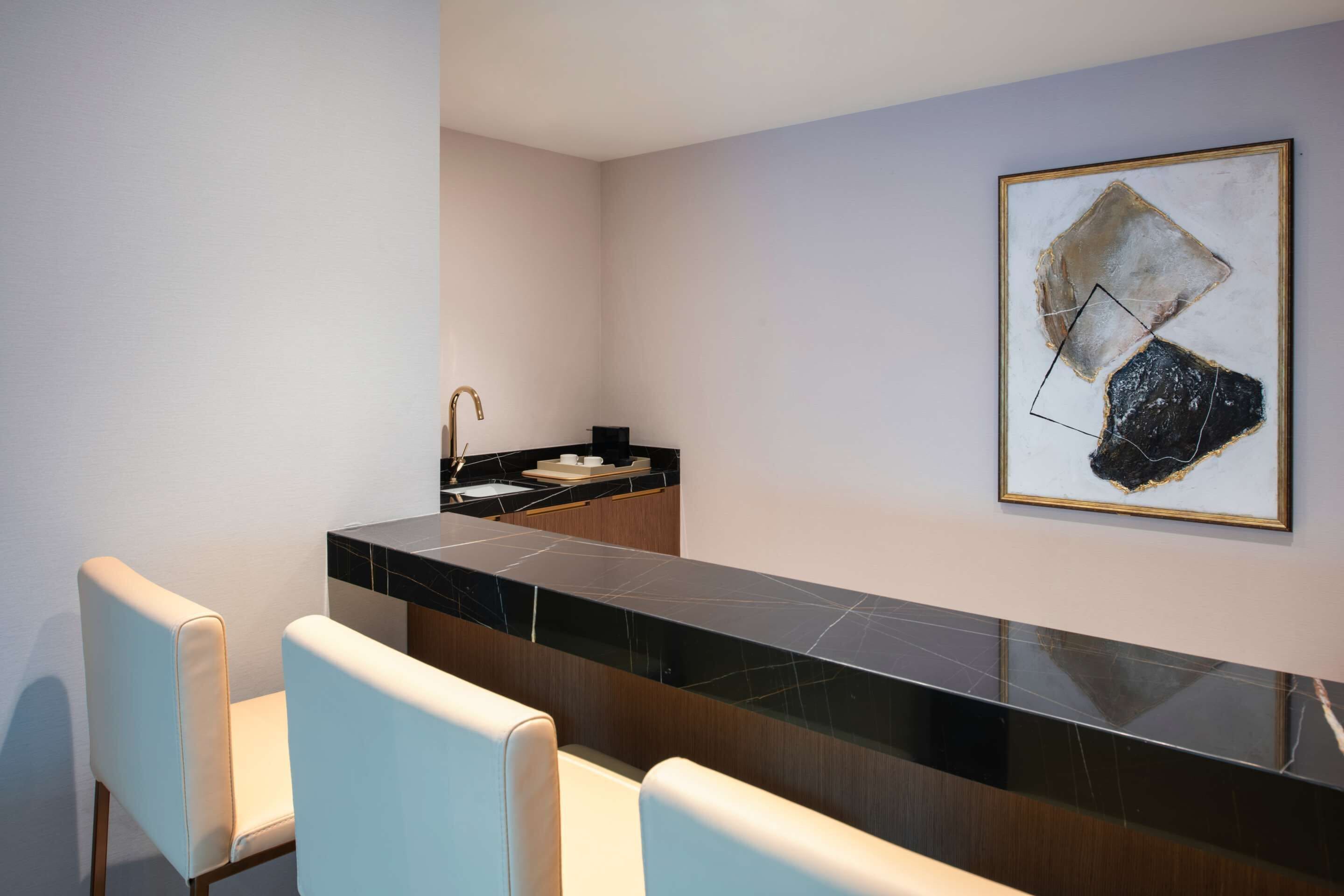 A modern kitchen with a marble countertop, a sink, and a painting on the wall. There are two white chairs in front of the counter.