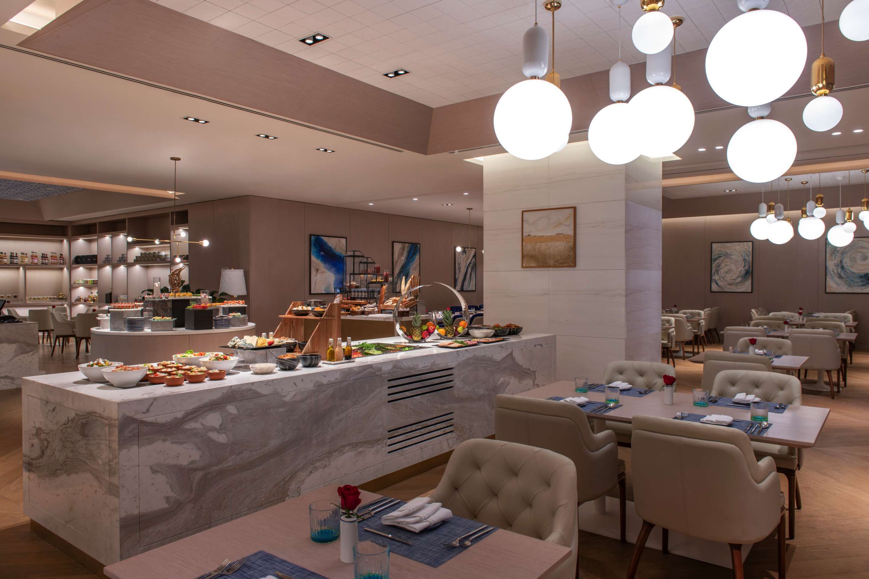 A modern hotel dining area with marble counters, round chandeliers, and neatly arranged tables and chairs.