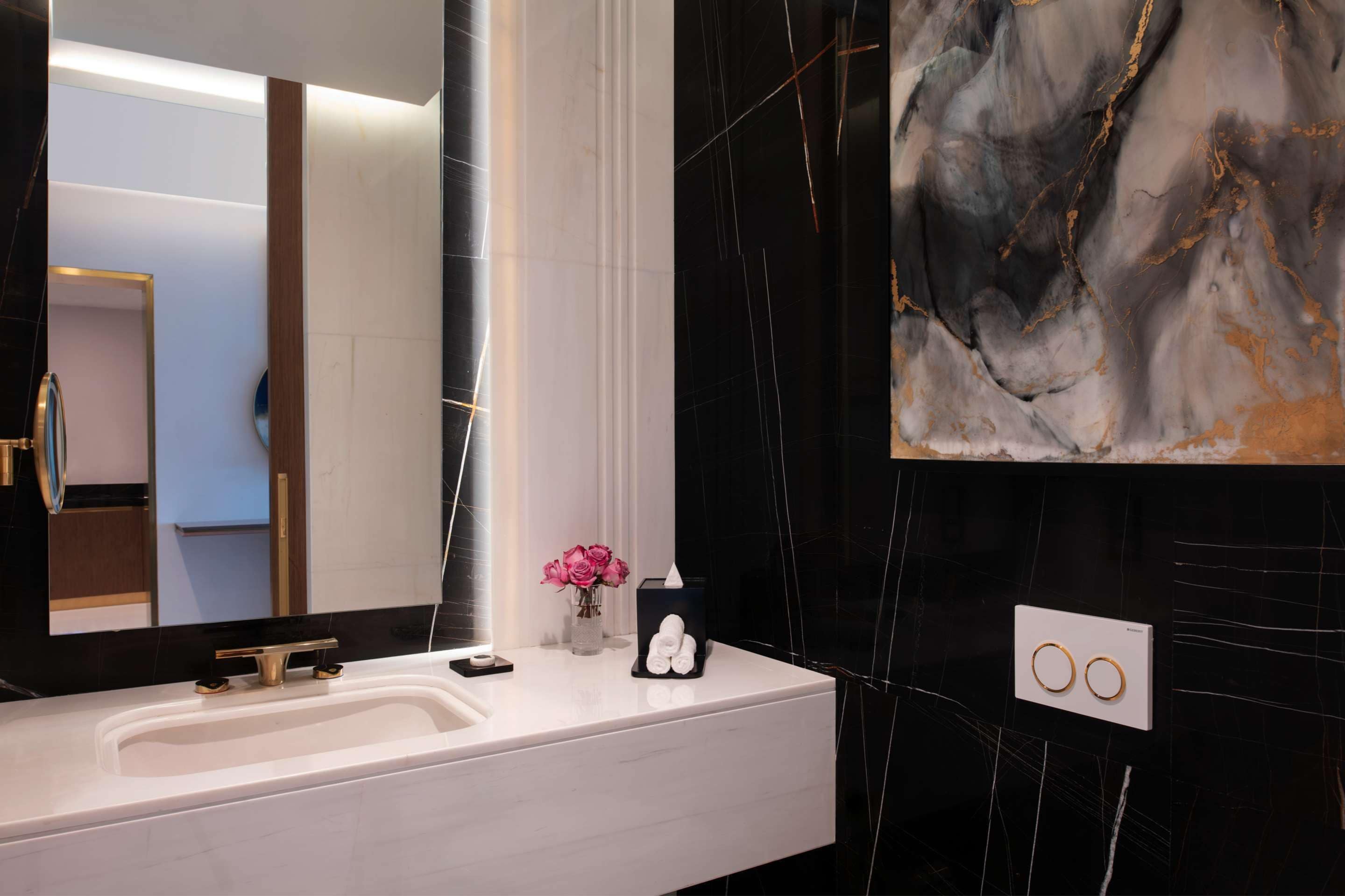 Un baño moderno con tocador blanco, un espejo grande, pared de mármol y un detalle decorativo sobre el lavabo.