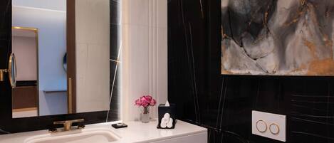 Un baño moderno con tocador blanco, un espejo grande, pared de mármol y un detalle decorativo sobre el lavabo.