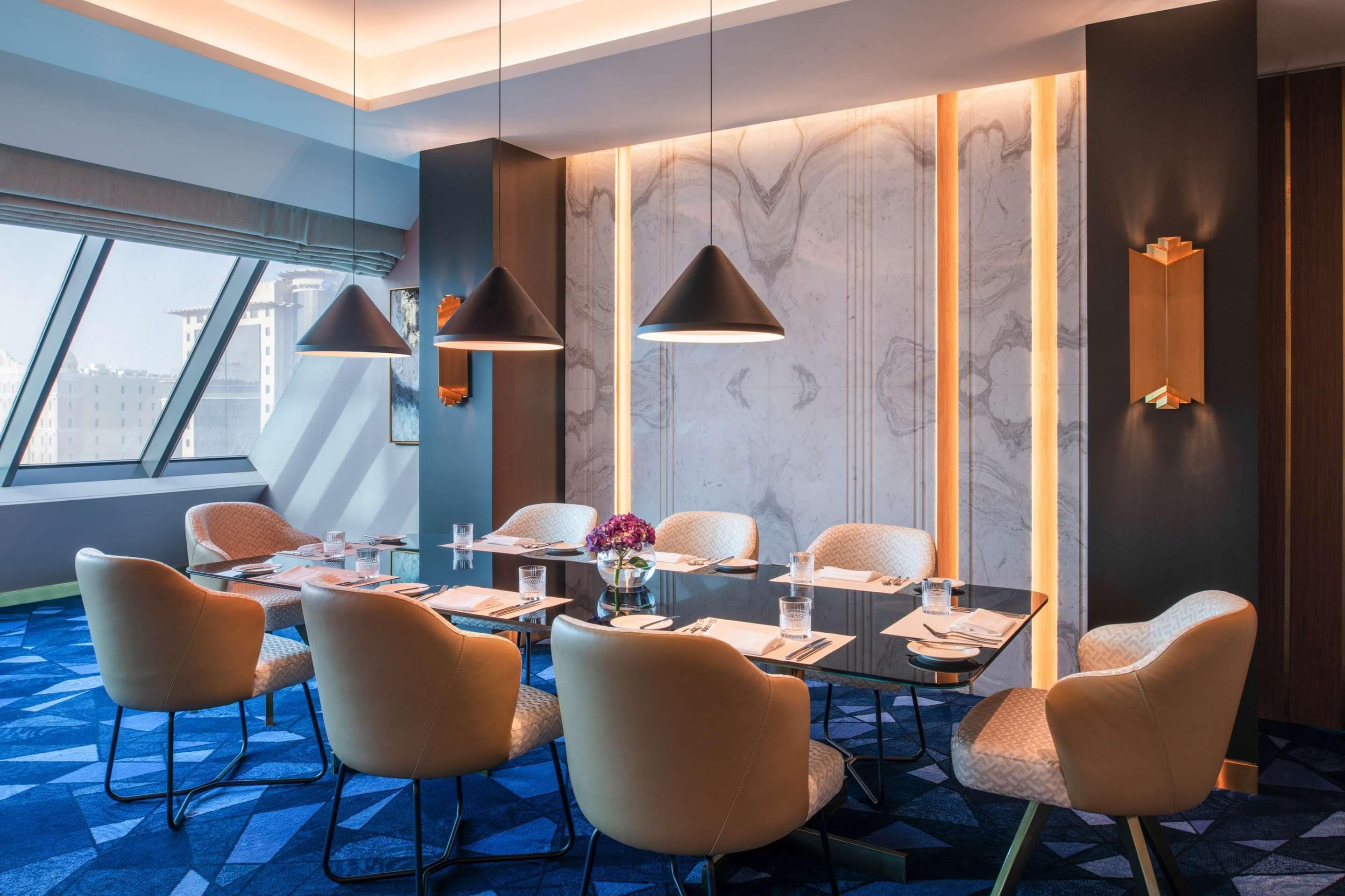 A modern hotel room with a dining area featuring a large table, comfortable chairs, and pendant lighting.