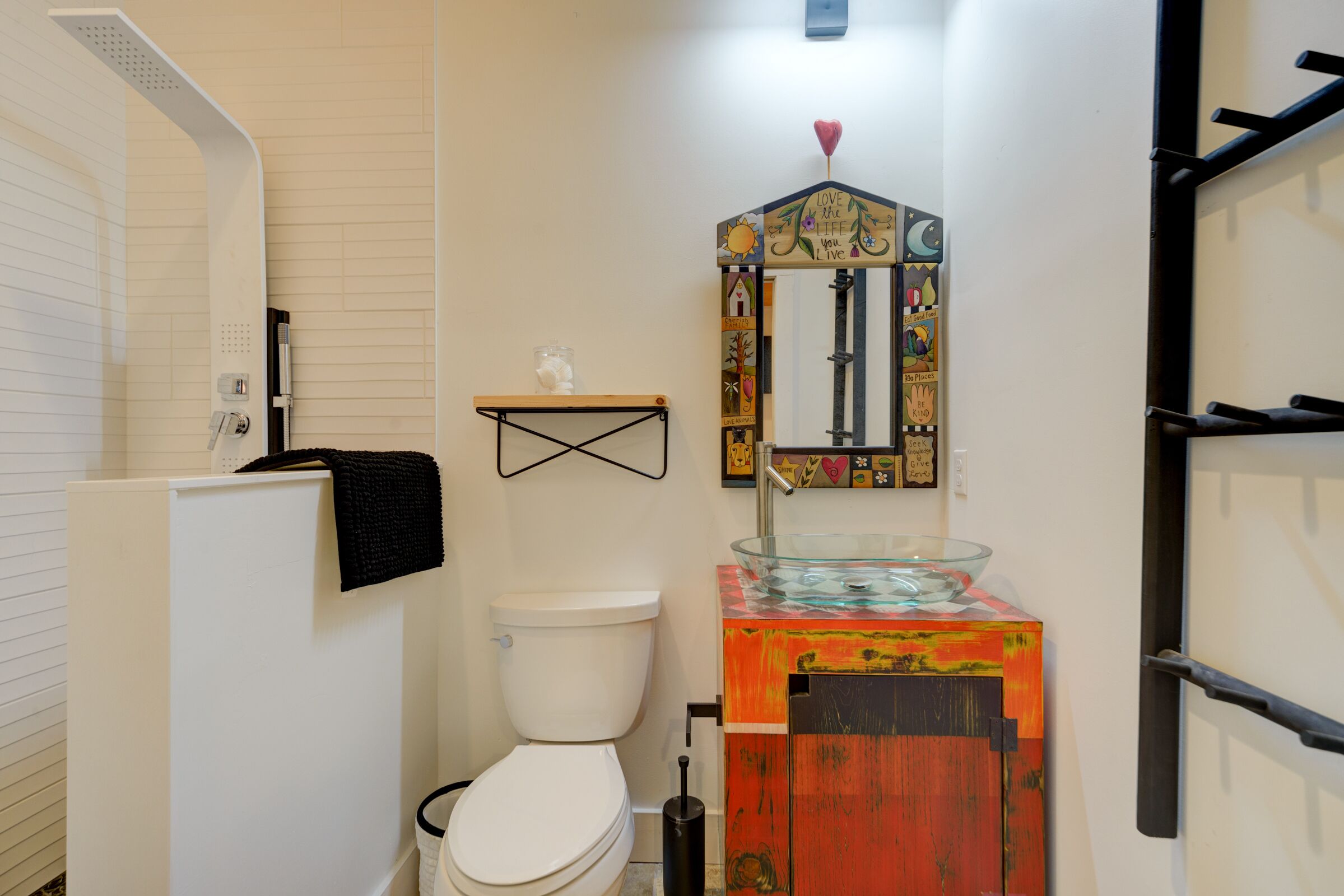 A bathroom with a white toilet, a glass sink with a colorful wooden base, a shelf with decorative items, and a shower area.