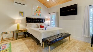 A bedroom with a wooden ceiling, a bed with a black headboard, a bench, a TV, and a window with curtains. - Charming 2BR Uptown Cottage - Artsy & Vibrant! (St. Augustine)
