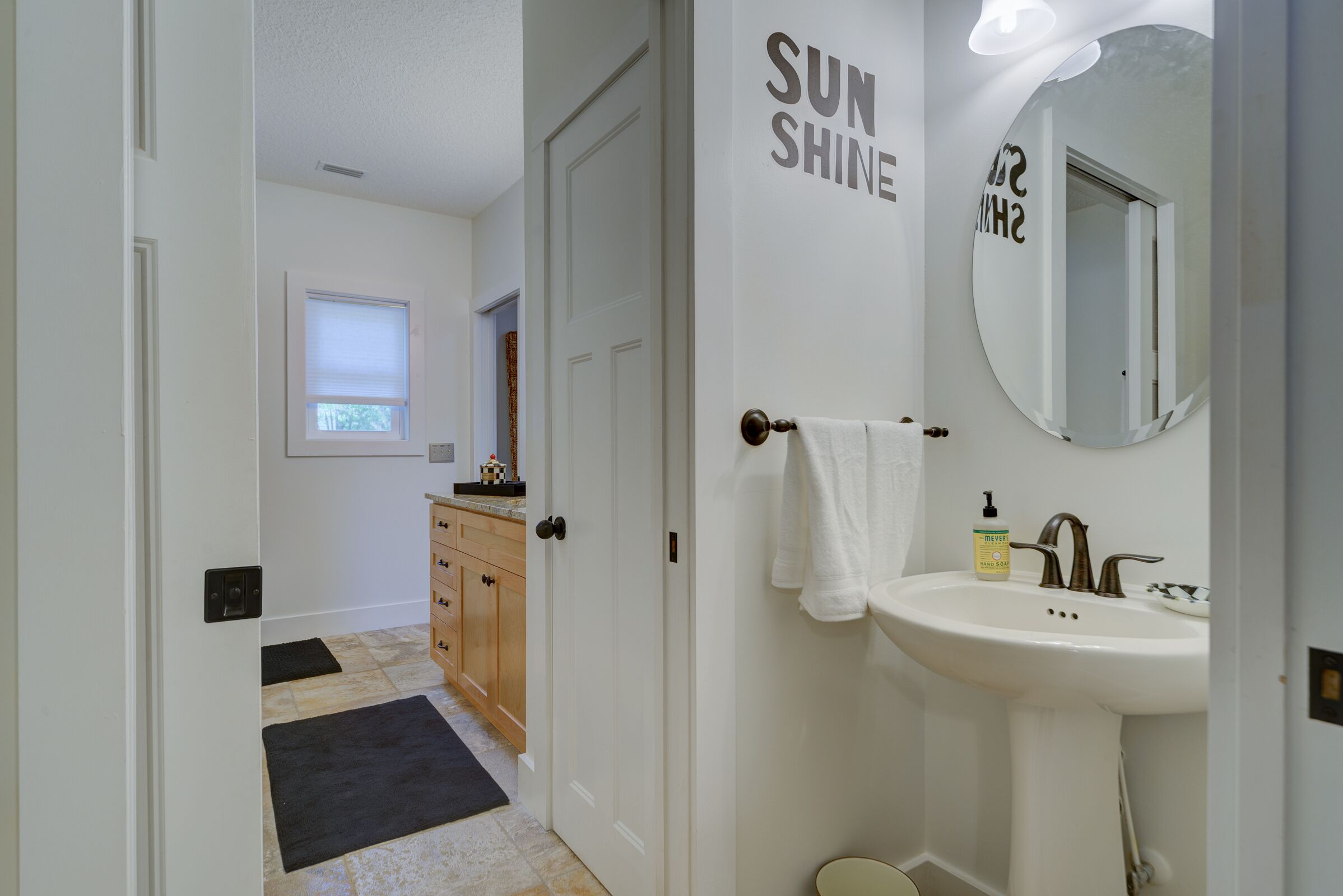 A bathroom with a white sink, a mirror, and a towel hanging on the door. There is a window with blinds and a door leading to another room.