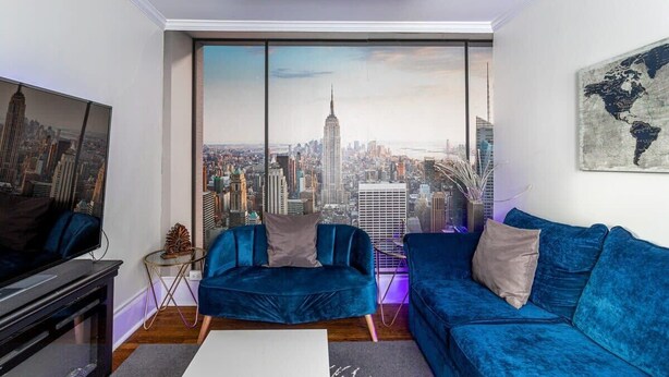 A modern living room with a large window offering a cityscape view, a blue sofa, a small side table, and a flat-screen TV. - The Fred Deluxe 7 BR by I95, Train, HarborVw, Stamford Beaches & Workspace

 (Stamford)
