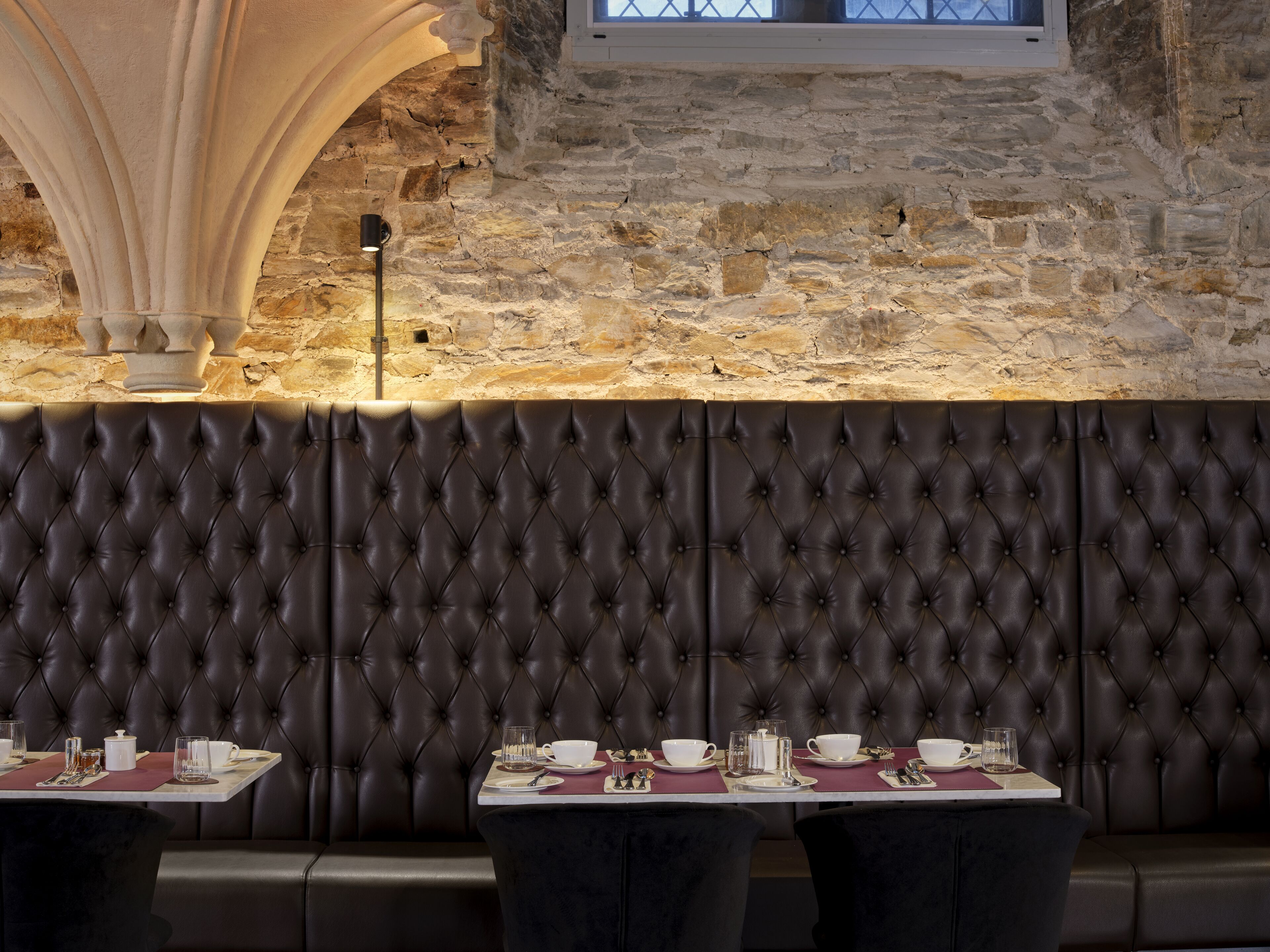 A restaurant with a stone wall, a dark upholstered booth seating area, and tables set for dining.