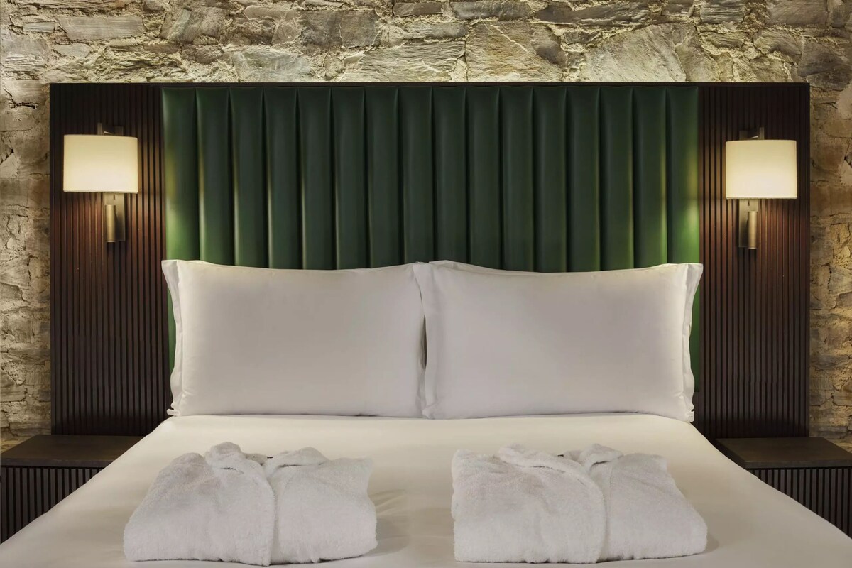 A hotel room with a stone wall, a green headboard, two white pillows, two white towels, and two wall-mounted lamps.