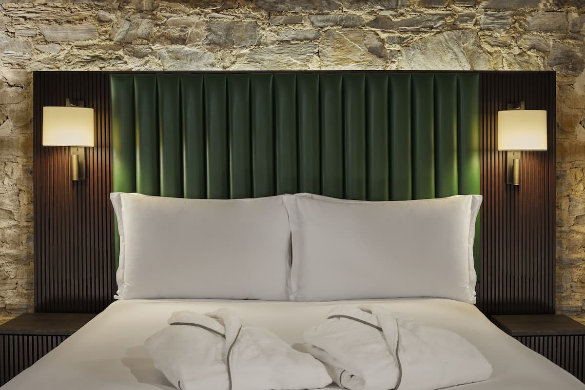 A bed with two pillows and two towels, flanked by two wall-mounted lamps, against a stone wall backdrop.