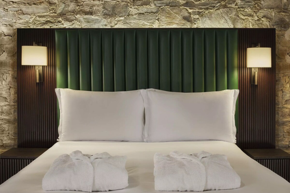 A hotel room with a stone wall, a green headboard, two white pillows, two white towels, and two wall-mounted lamps.