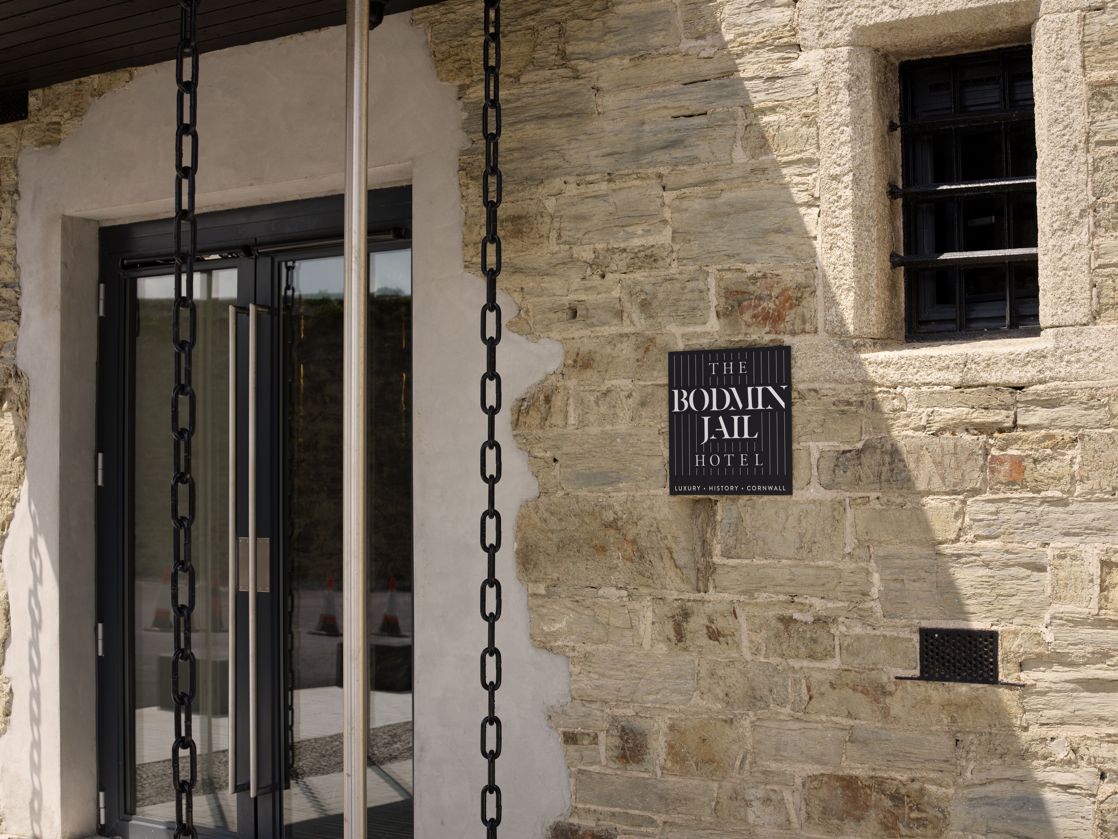 A stone building with a sign for The Bodmin Jail Hotel, featuring a chain-link security gate and a window.