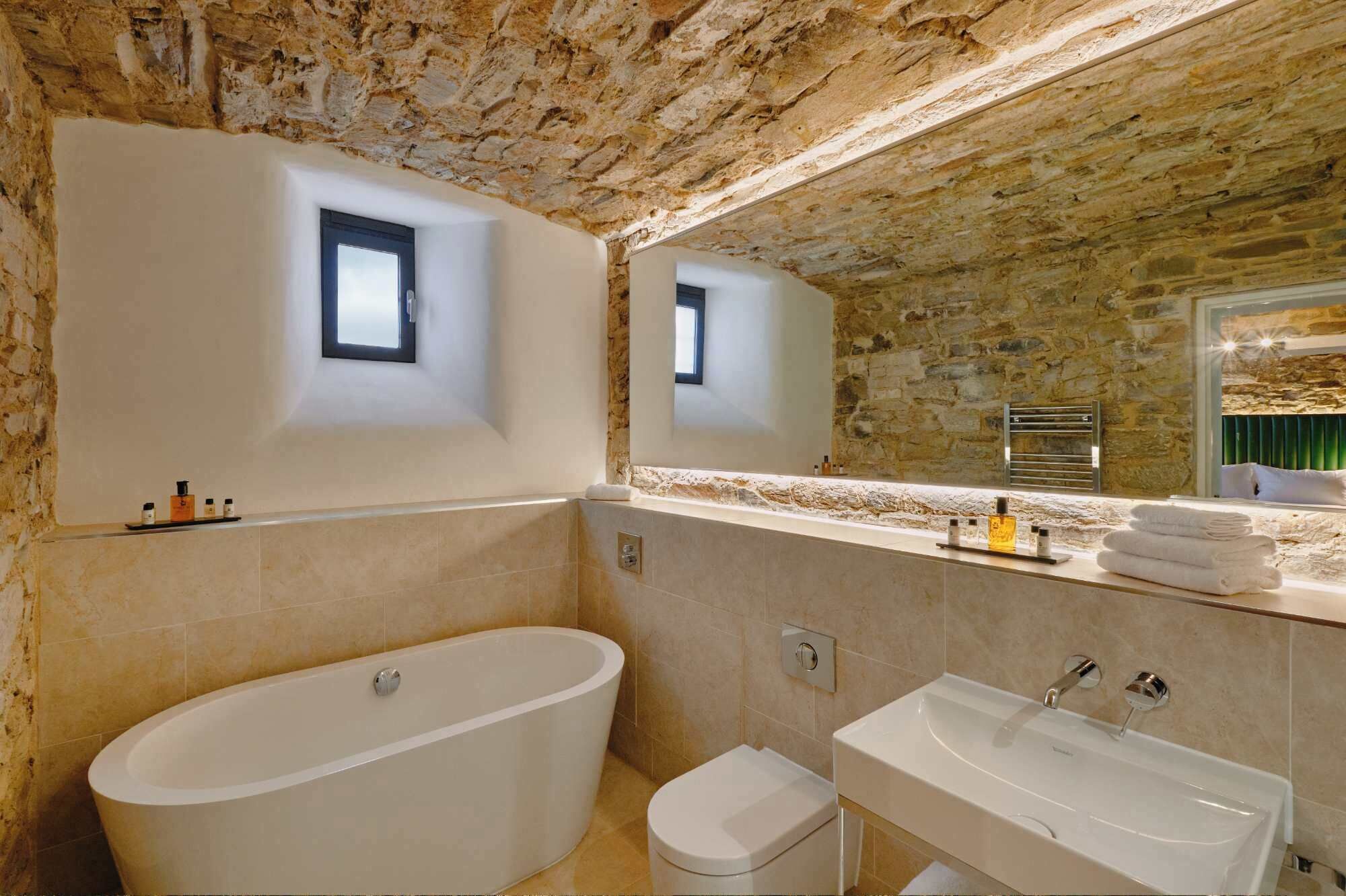 A bathroom with a bathtub, toilet, and sink, stone walls, and a small window.