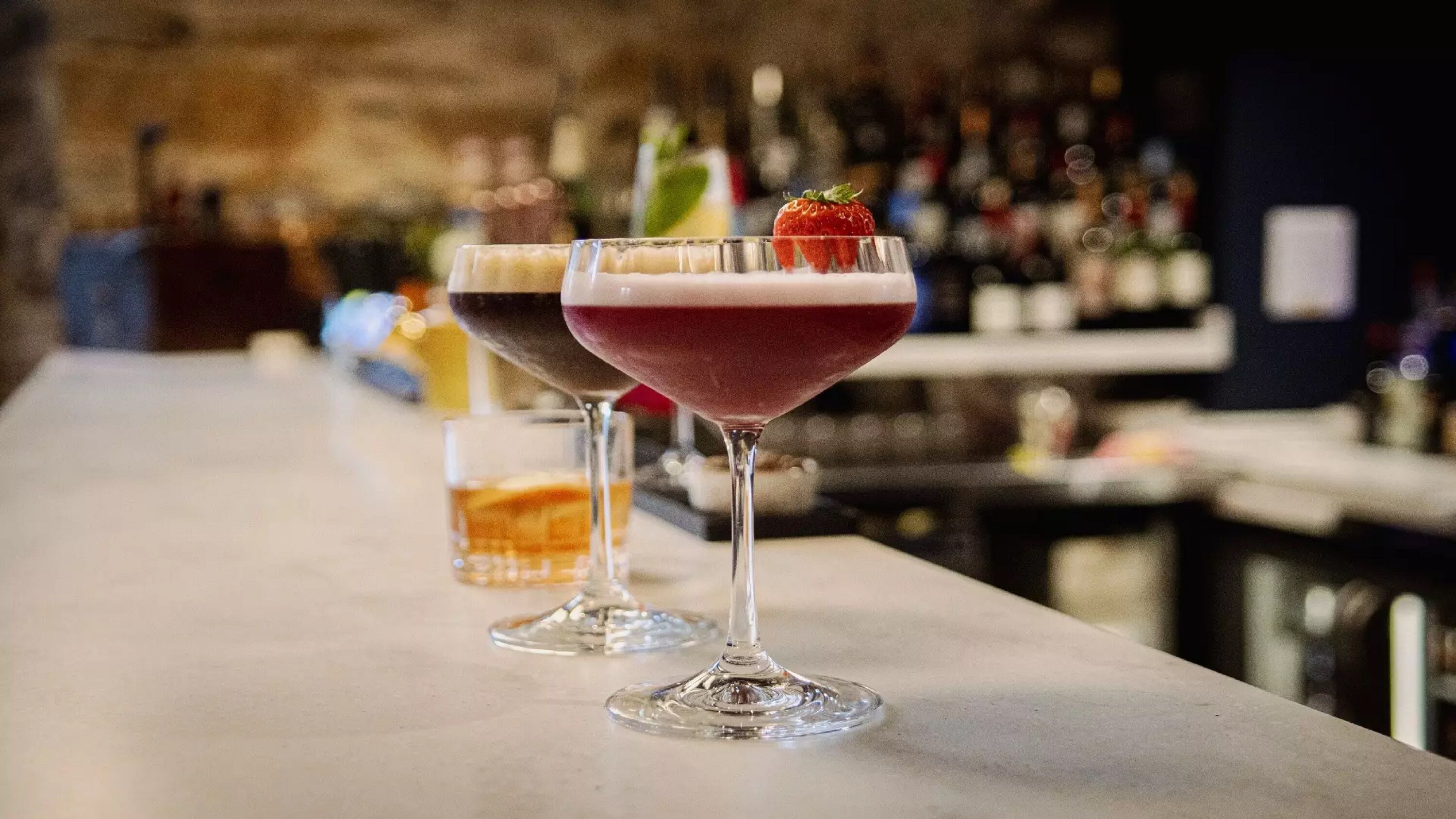 Three cocktails on a bar counter with a blurred background of bottles.