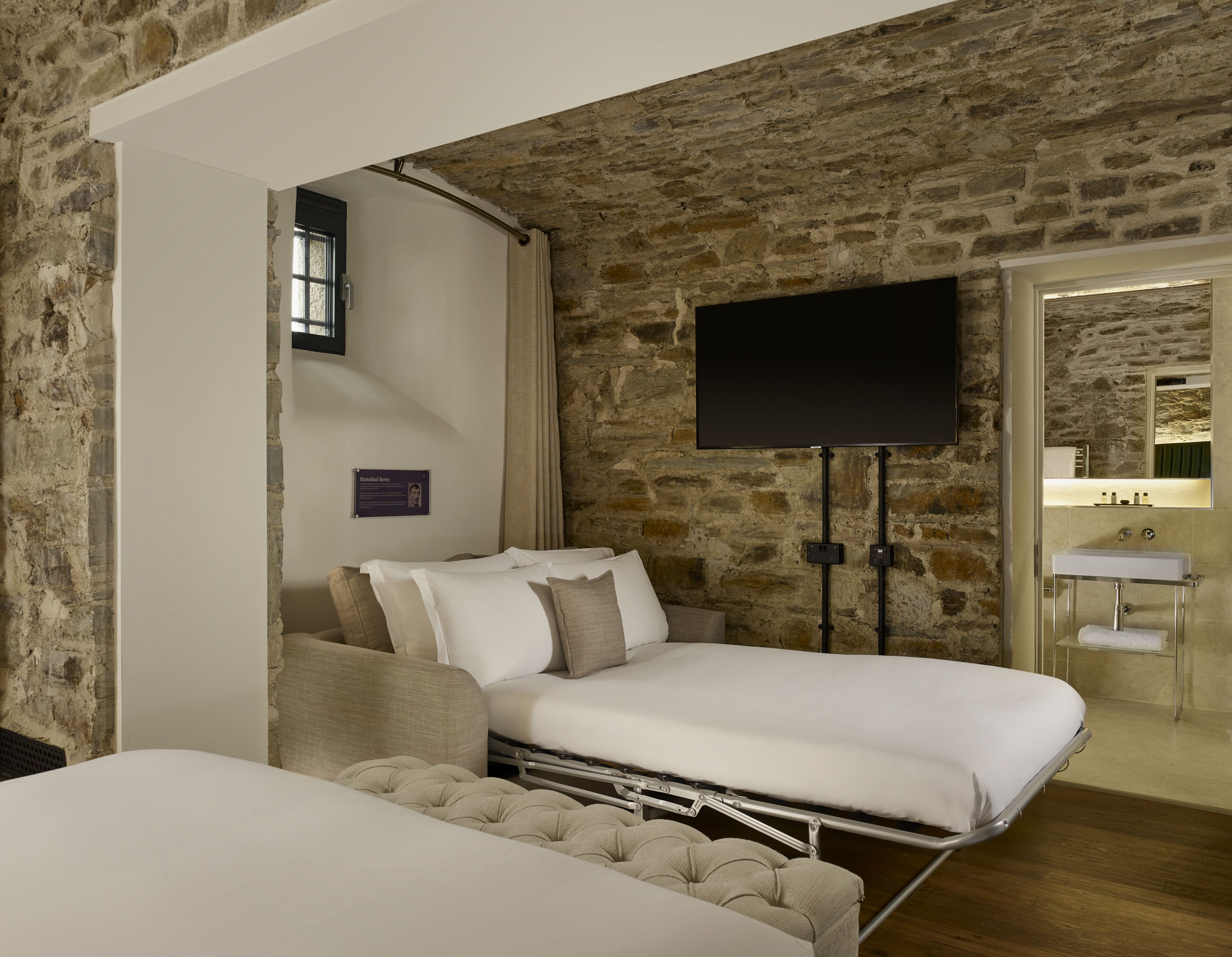 Una camera d'albergo con parete in pietra, un letto, una televisione e un bagno con un lavandino.
