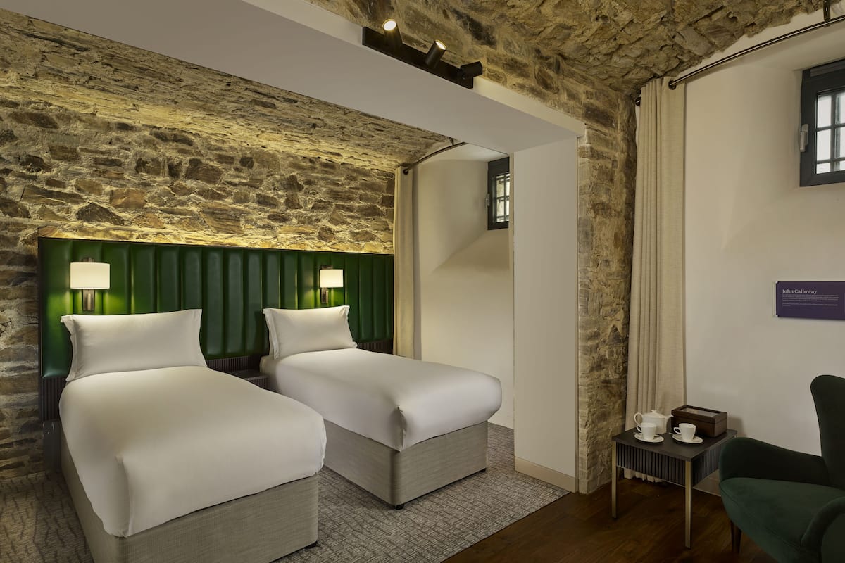 A hotel room with two beds, a green headboard, a stone wall, a small table with a teapot, and a green armchair.