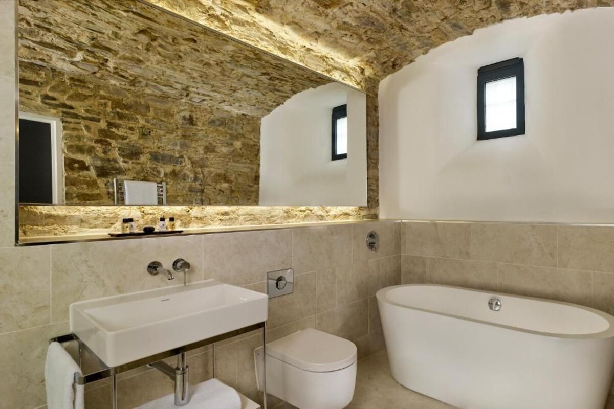 A bathroom with a stone wall, a large mirror, a sink, a toilet, and a bathtub.
