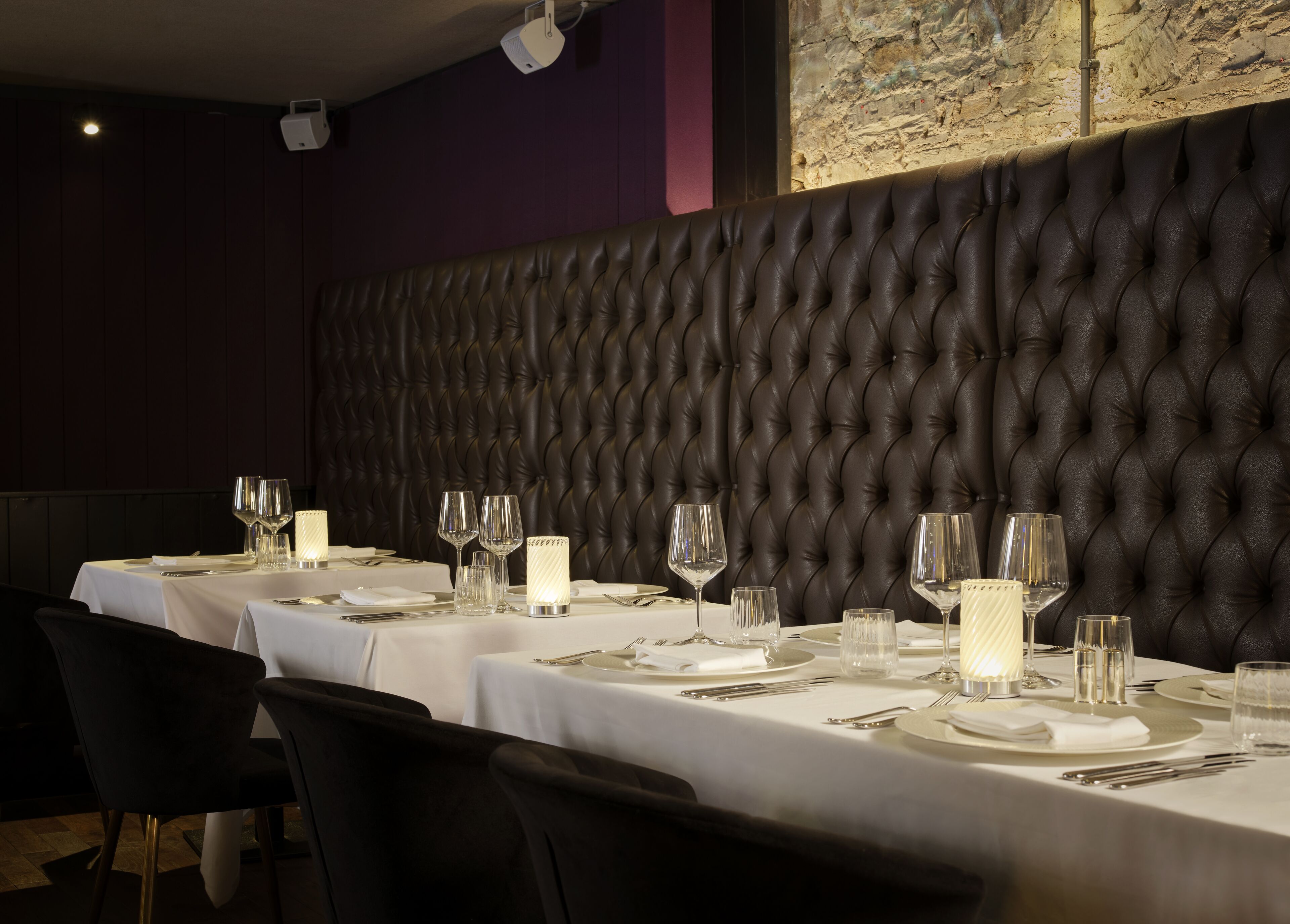 Un espace repas avec une longue table dressée pour un repas, agrémenté d’un mur en cuir capitonné et d’un mur en pierre orné de graffitis.