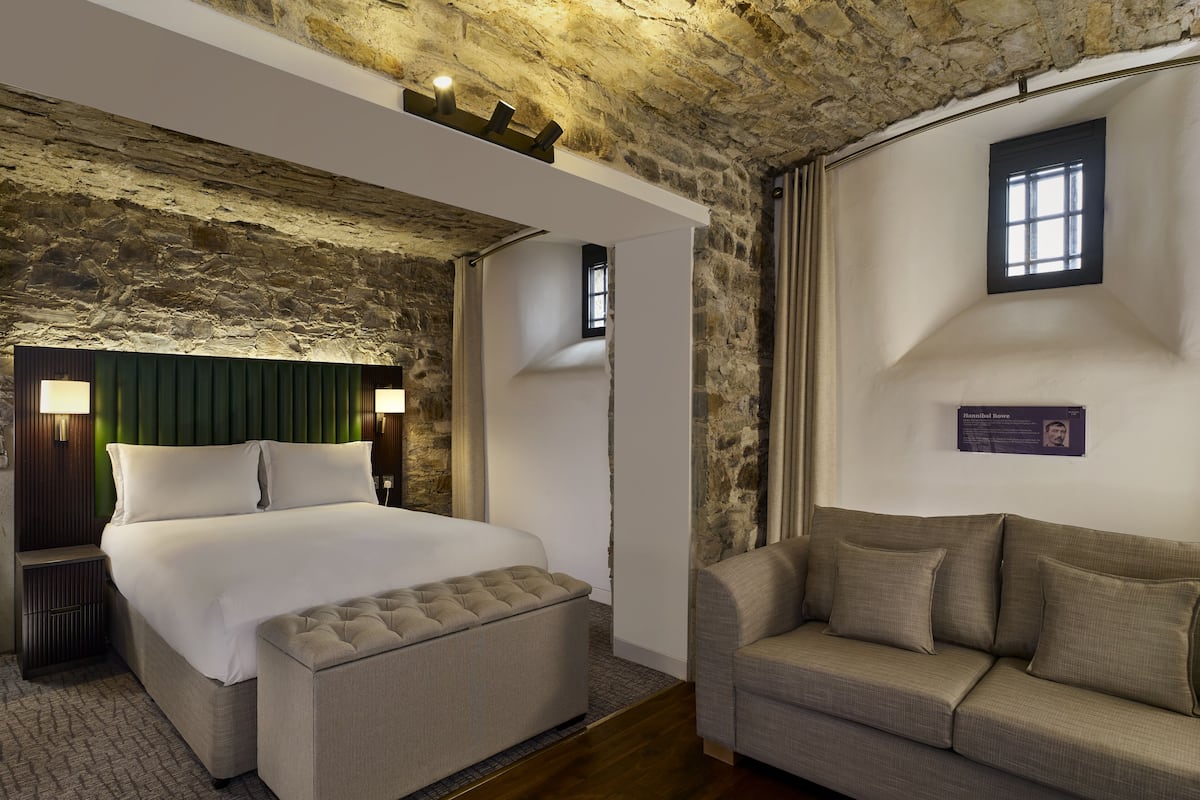 A bedroom with a stone wall, a bed with a green headboard, a bedside table, a nightstand, a sofa, and a small window.