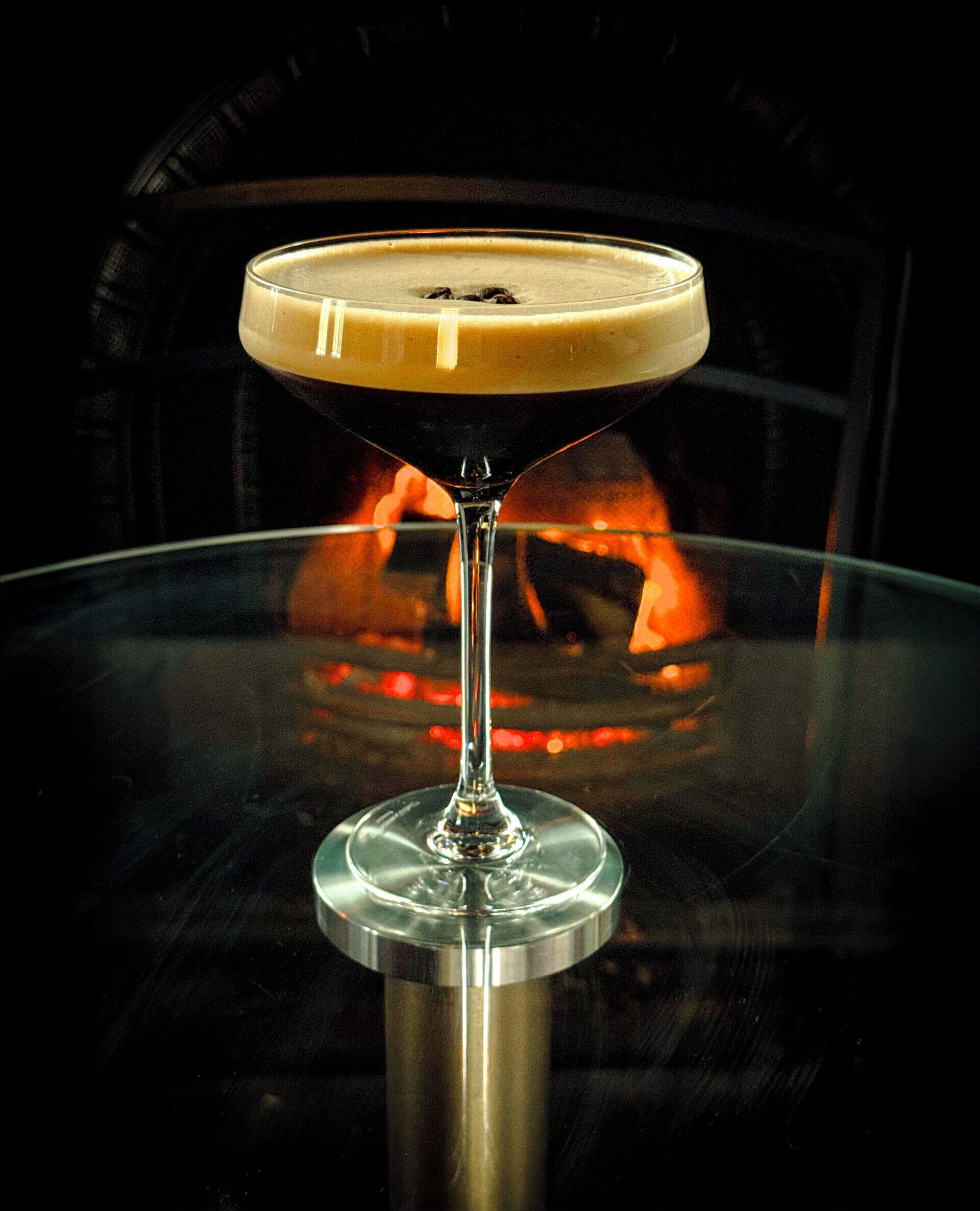 Ett martiniglas med en skummande drink på ett genomskinligt glasbord, med en brasa i bakgrunden.