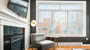 Un salon moderne avec une cheminée, un téléviseur à écran plat fixé au-dessus, un canapé en cuir gris, une petite table ronde et une grande fenêtre avec des stores.