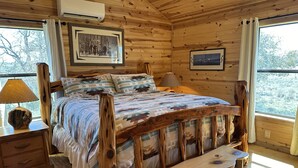 A wooden cabin bedroom with a rustic bed, a bedside lamp, and a view of the outdoors. - Arrowhead Cabin at Wolf Creek Guest Ranch (Fredericksburg)