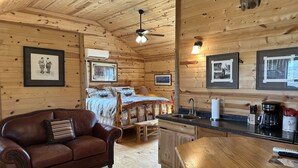 A cozy cabin interior with a wooden ceiling, a bed with patterned bedding, a leather sofa, a dining table, and a kitchen area with a sink and coffee maker. - Arrowhead Cabin at Wolf Creek Guest Ranch (Fredericksburg)