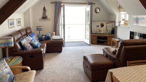 A spacious loft-style living room with a high ceiling, a large sliding glass door leading to a balcony, and a cozy seating area with a brown leather sofa and ottoman.