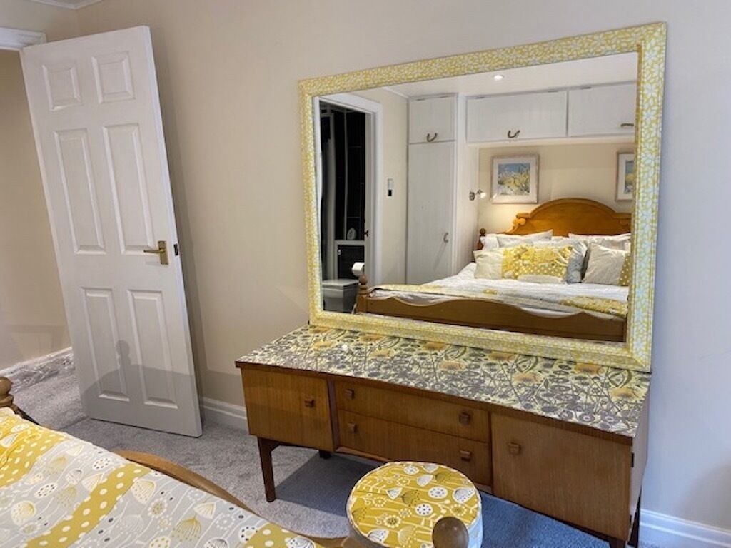 A bedroom with a large mirror, a bed with patterned bedding, a wooden dresser, and a small stool.