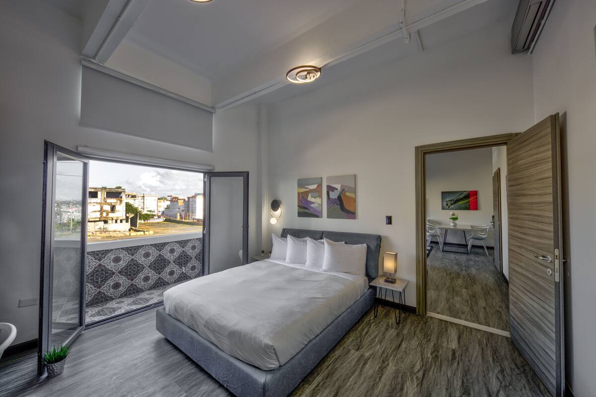 A modern bedroom with a large bed, a small bedside table, a mirror, and a view of the outside.