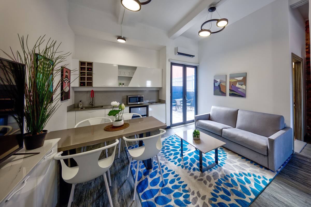 A modern living room with a grey sofa, a coffee table, and a dining area with a white table and chairs. There is a kitchen with white cabinets and a microwave.