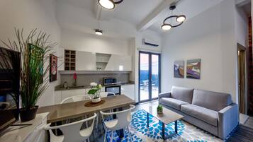 Uma sala de estar moderna com um sofá cinza, uma mesa de centro e uma área de jantar com uma mesa e cadeiras brancas. Há uma cozinha com armários brancos e um micro-ondas.