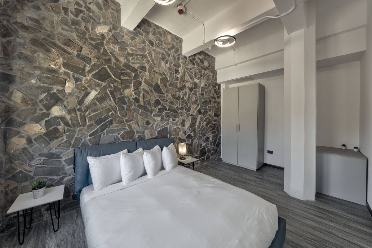 A modern bedroom with a stone wall, a bed with white bedding, a bedside table with a lamp, and a wardrobe.