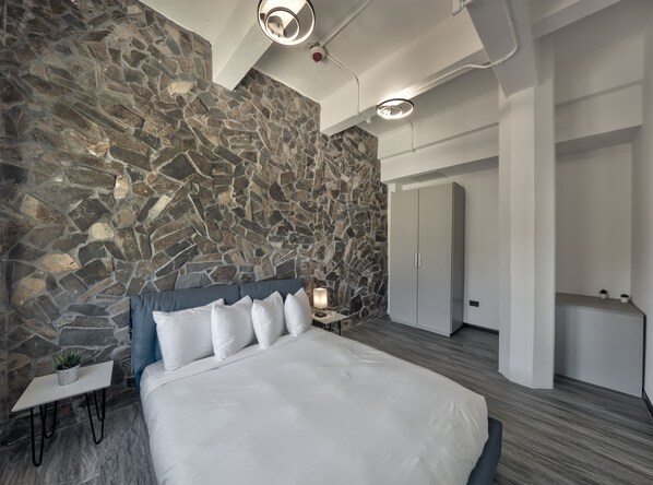 A modern bedroom with a stone wall, a bed with white bedding, a bedside table with a lamp, and a wardrobe. - Hotel Puerto Valdes (San Juan)