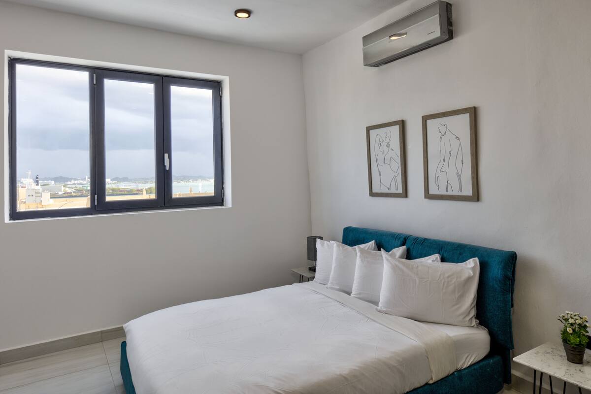 A bedroom with a large window, a bed with white bedding, a blue headboard, and two framed artworks on the wall.