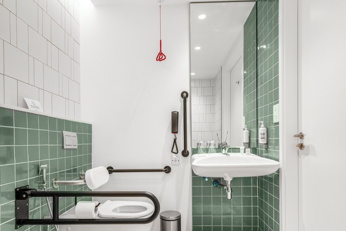 Un bagno moderno con un lavandino bianco, una vasca da bagno con maniglia di sostegno e pareti rivestite di piastrelle verdi.