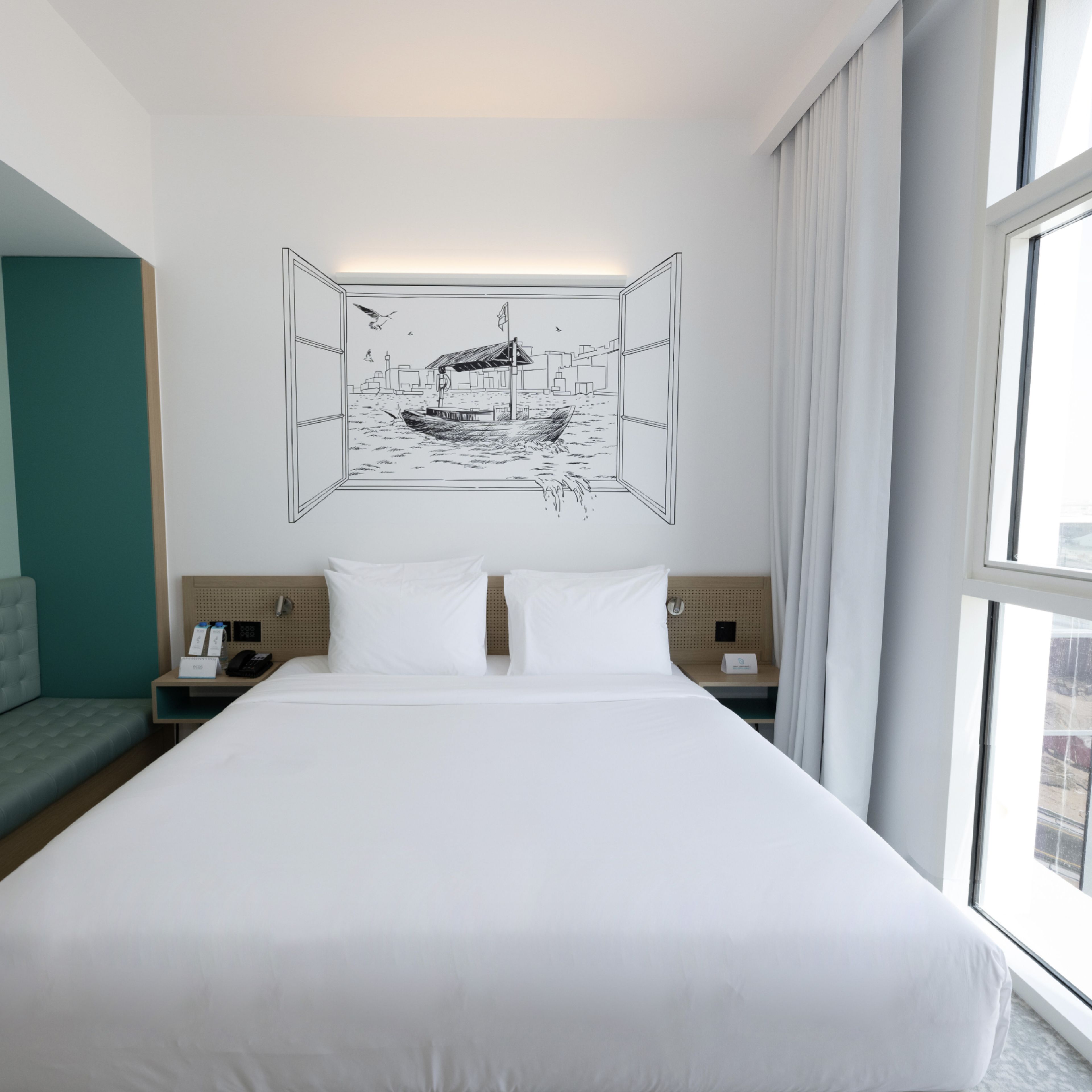 A hotel room with a large bed, a wall-mounted artwork of a boat, a desk with a phone, and a window with curtains.