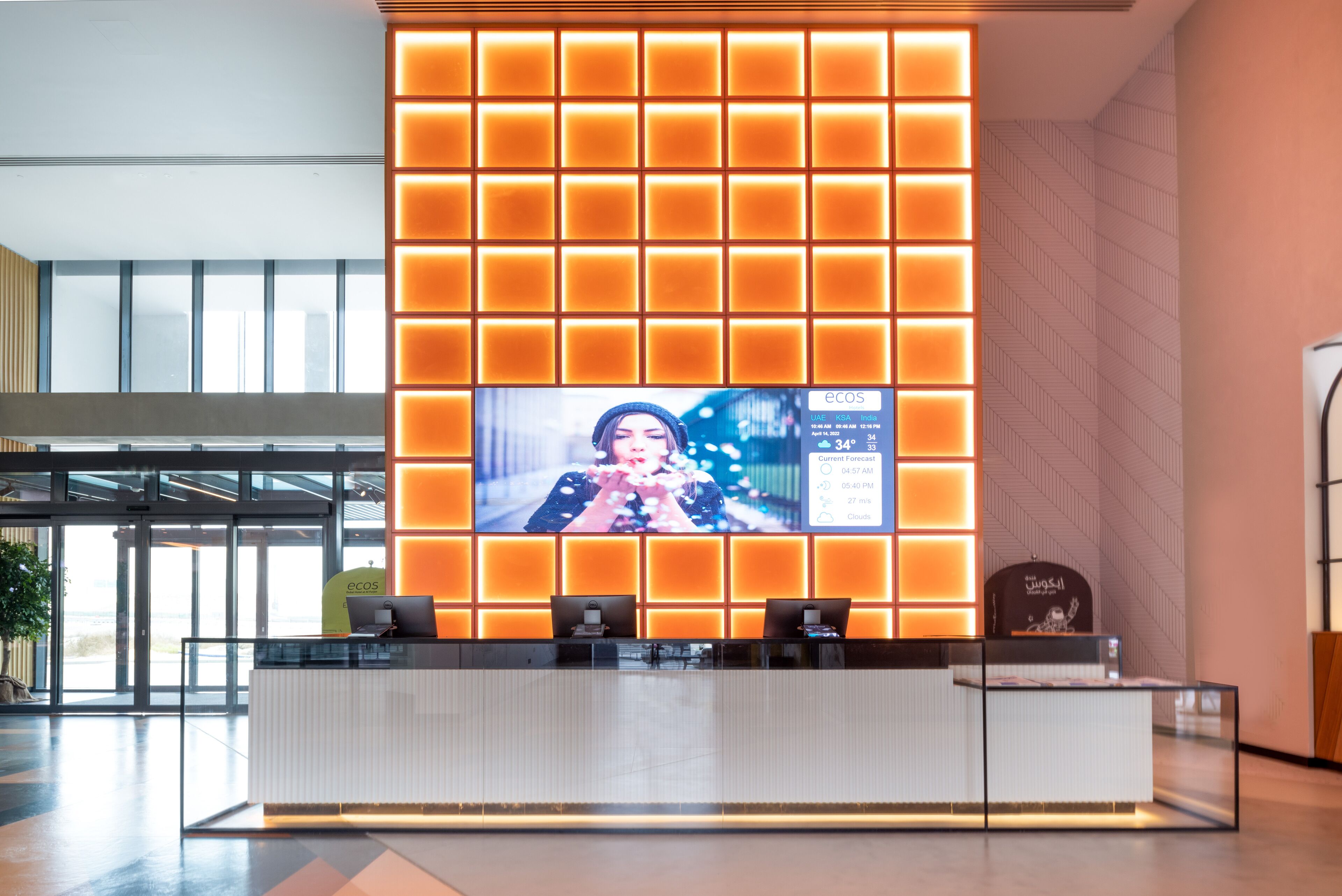 A reception desk with a large screen displaying weather information, a modern design with orange lighting, and a view of the lobby area.