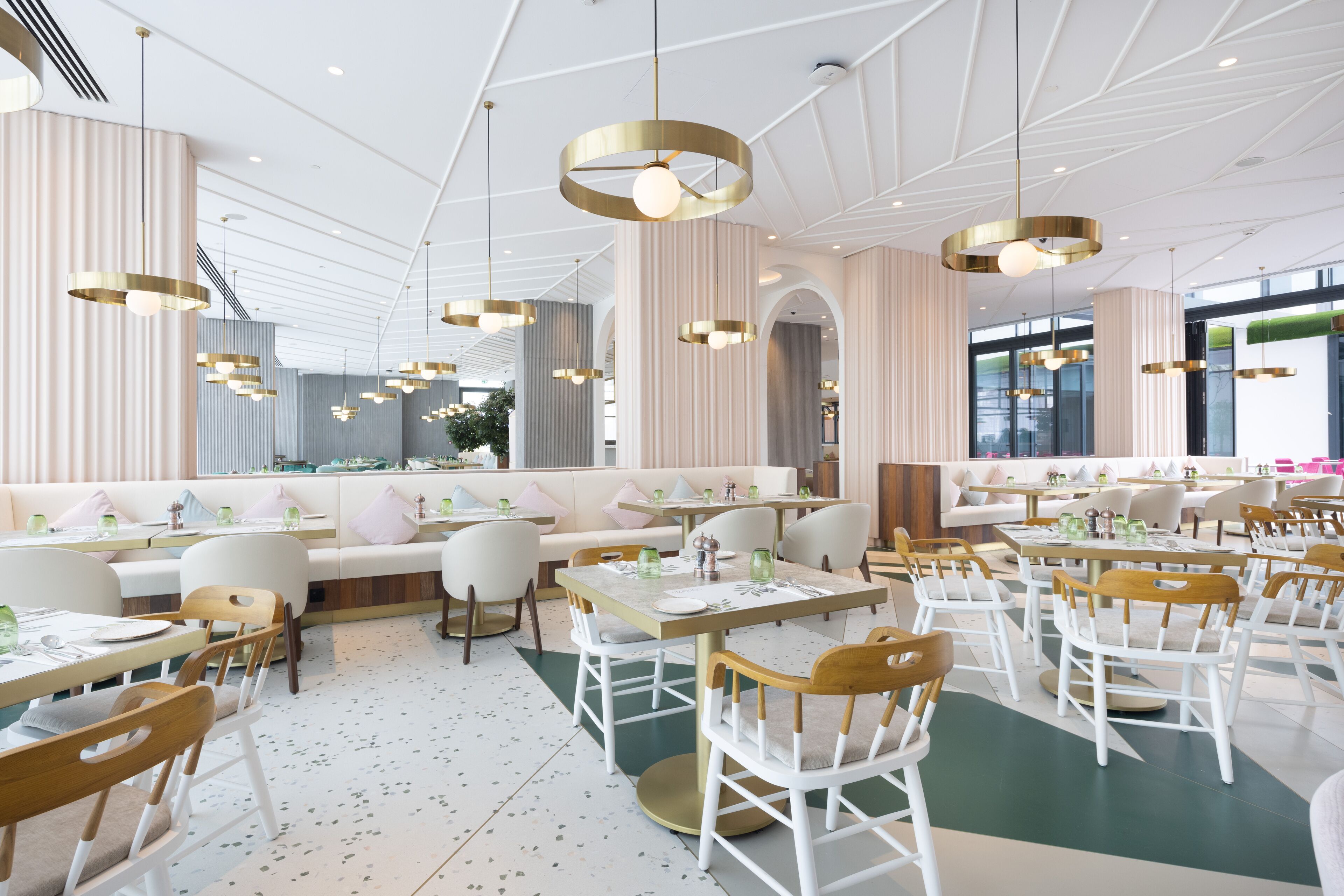 Un restaurante moderno con decoración en blanco y beige, con mesas redondas, sillas de madera e iluminación colgante.