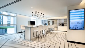 Een moderne hotellobby met een centraal gelegen bar, een digitaal scherm met conferentietijden en een stadszicht door grote ramen.
